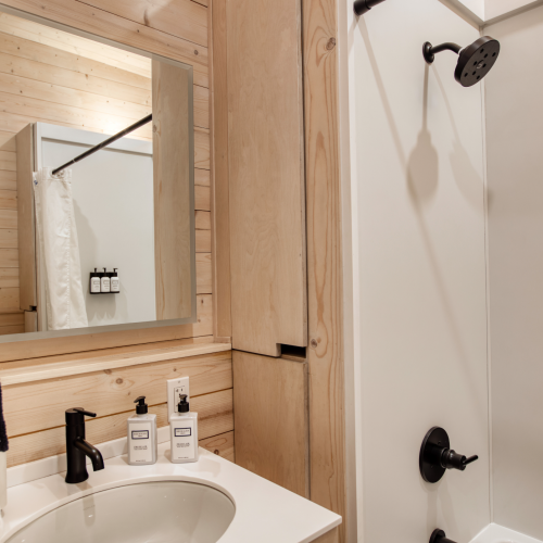 A small bathroom with a wooden interior, featuring a sink, mirror, toiletries, and a shower area with a curtain and black showerhead.