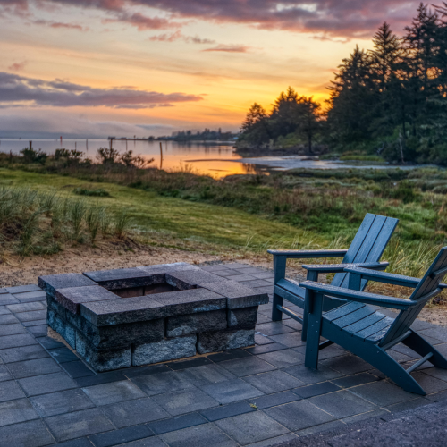 A scenic sunset view with two chairs by a stone fire pit, overlooking grass, trees, and a calm water body under a colorful sky.