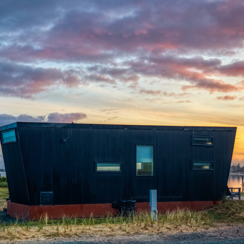 A modern, uniquely shaped black house near a body of water, with a colorful sunset sky in the background.