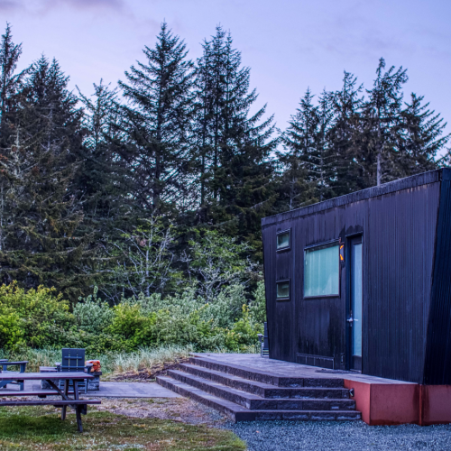 A modern tiny house near a forest with a picnic table and chairs on a grassy area under a clear sky, featuring steps leading to the entrance.