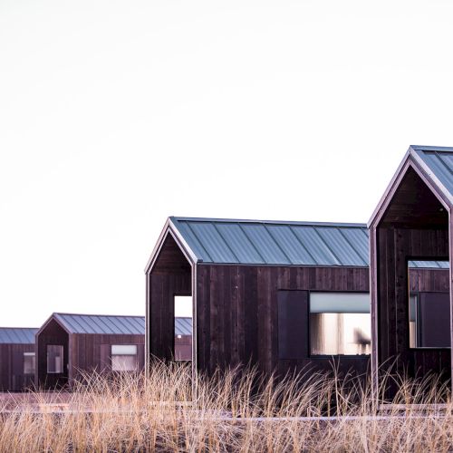 The image shows a series of small, modern wooden cabins with pitched roofs, set against a dry, grassy landscape under a clear sky.