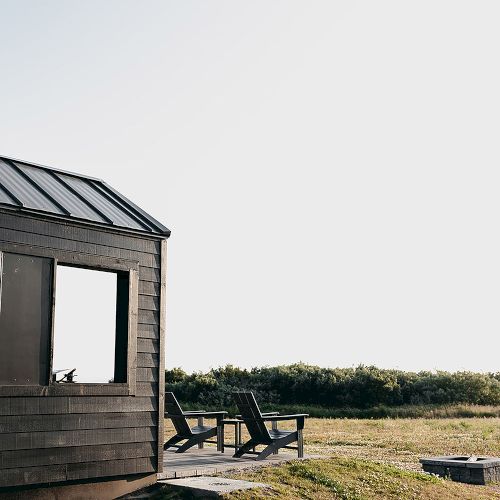 A wooden cabin with large windows is situated on a grassy field, with two chairs outside overlooking a scenic landscape.