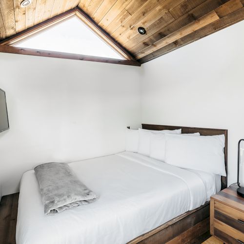 A small bedroom with a white bed, wooden ceiling, TV on the wall, window with a view, and a bedside table with a lamp and tissue box.