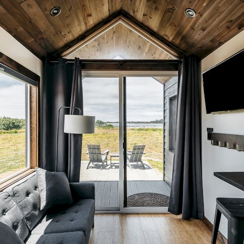 A cozy cabin interior with a couch, TV, and large windows showing a scenic view. A sliding door leads to a deck with outdoor seating.