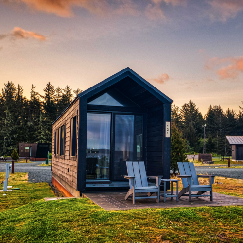 A small modern cabin with large windows surrounded by a grassy area and trees, featuring two chairs and a table outside.