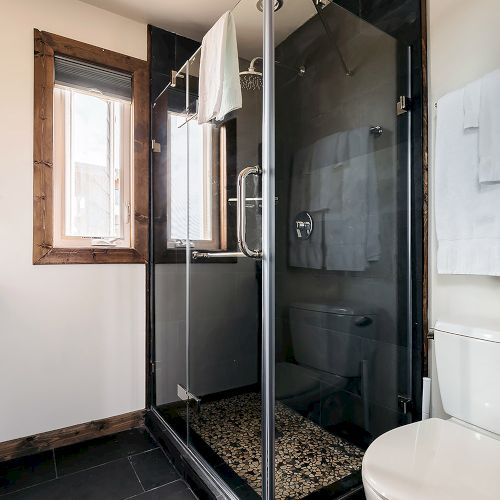 The image shows a modern bathroom with a glass shower enclosure, a window, towel rack, and a toilet on dark tile flooring.