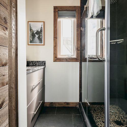 The image shows a small bathroom with a wooden wall, a sink with cabinets, a framed picture, and a glass-enclosed shower.