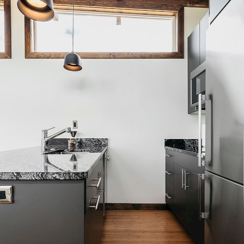 A modern kitchen with sleek gray cabinets, granite countertops, wooden flooring, hanging lights, and high ceilings with exposed beams.