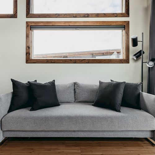 A modern living room with a gray sofa, black pillows, framed art, and large windows. Wooden flooring complements the contemporary décor.