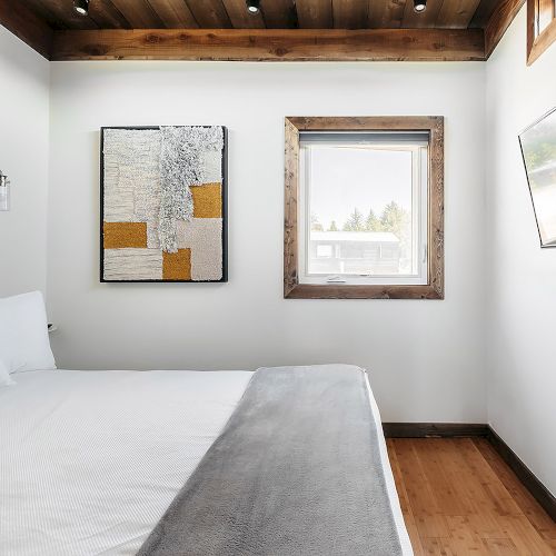 A minimalist bedroom features a bed, wall art, a window with a view, and a mounted TV on wooden floors.