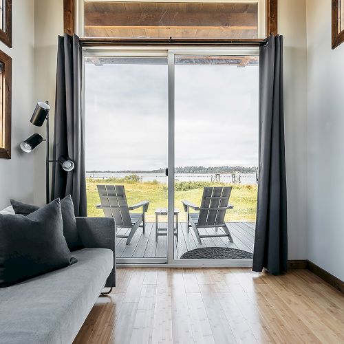 The image shows a cozy room with a gray sofa, wooden floor, and sliding glass doors leading to an outdoor area with two chairs facing a scenic view.