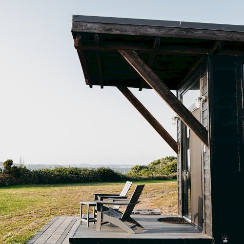 A modern cabin with a wooden deck and two chairs overlooks a grassy field and distant water under a clear sky.