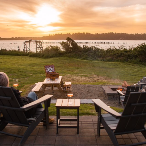 A serene sunset view over a lake, with two chairs facing the water, a firepit, and a table in a grassy area.