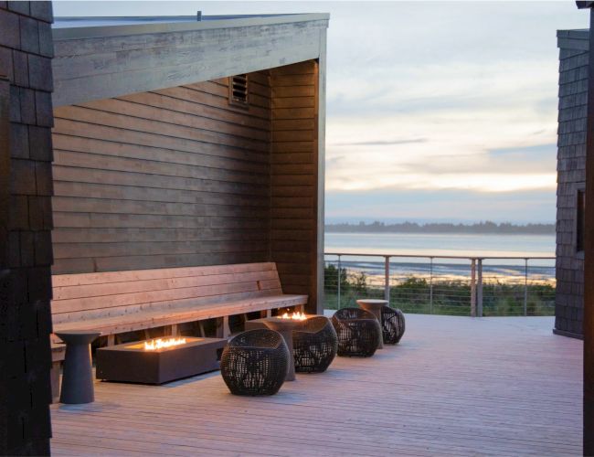 An outdoor seating area with a wooden bench, fire pits, and a view of the water at sunset.