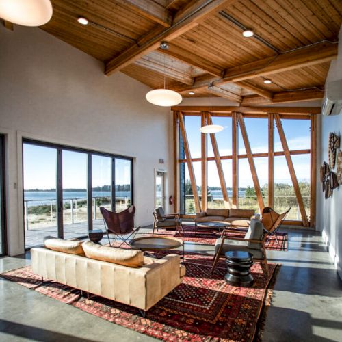 A modern living room with wooden ceilings, large windows, sofas, chairs, and rugs, offering a scenic outdoor view, has cozy lighting.