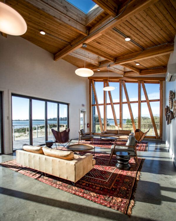 A modern living room with wooden ceilings, large windows, sofas, chairs, and rugs, offering a scenic outdoor view, has cozy lighting.