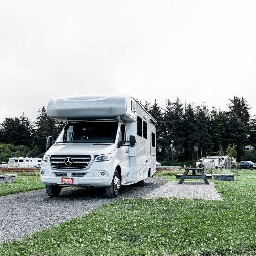 The image shows a white motorhome parked on a camping site with trees in the background.