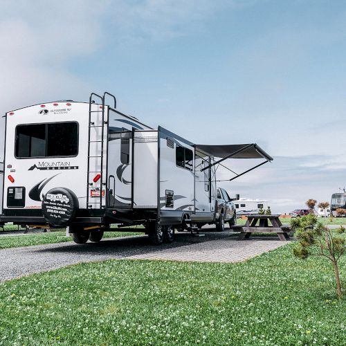 An RV parked on a gravel path in a grassy area with a picnic table and a small tree nearby, under a partly cloudy sky.