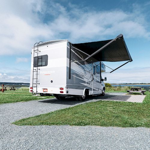 A parked RV with an extended awning sits on a gravel path by a grassy area and picnic table, under a blue sky with clouds.