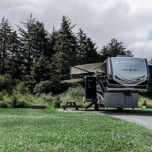 The image shows a parked RV with slide-outs extended in a grassy area surrounded by trees, with a clear sky in the background.