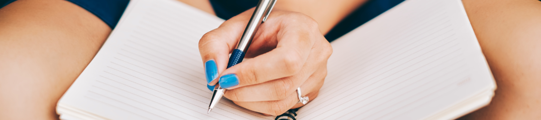 A person writing in a spiral-bound notebook with a pen, wearing blue nail polish and seated with the notebook on their lap.