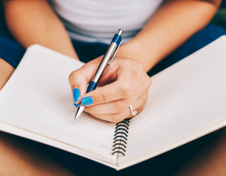 A person writing in a spiral-bound notebook with a pen, wearing blue nail polish and seated with the notebook on their lap.