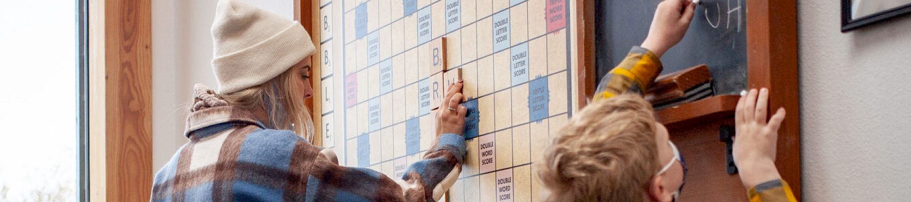 Three people, including two kids and an adult, play on a large wall-mounted Scrabble board.