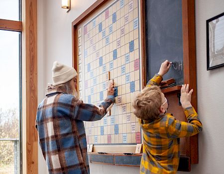 Three people, including two kids and an adult, play on a large wall-mounted Scrabble board.