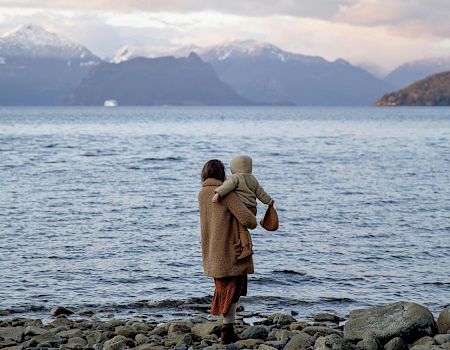 A person holds a child while standing on a rocky shore, gazing at a calm body of water with mountains in the background.