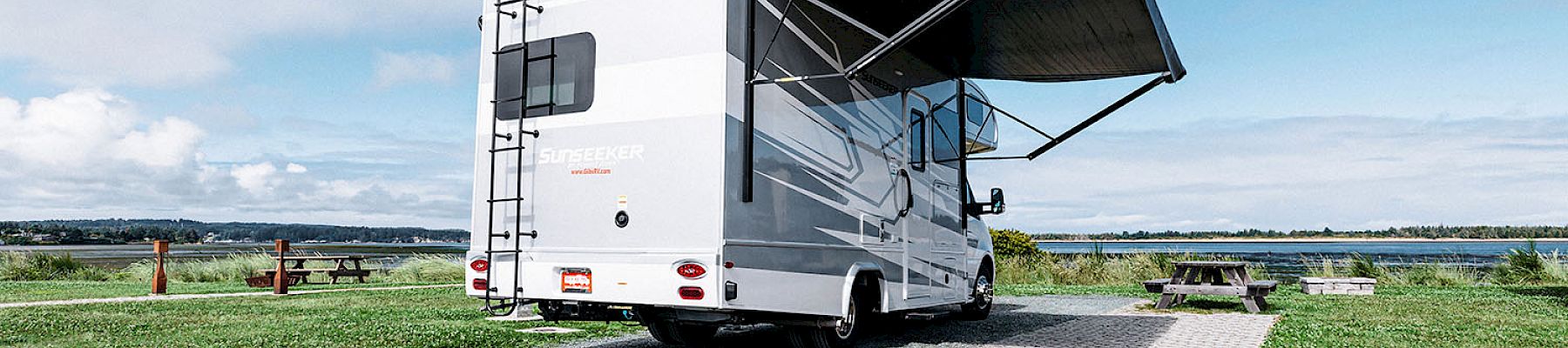 A parked RV with an extended awning, situated in a grassy area with a distant view of water and a cloudy sky.