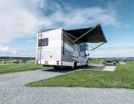 A parked RV with an extended awning, situated in a grassy area with a distant view of water and a cloudy sky.