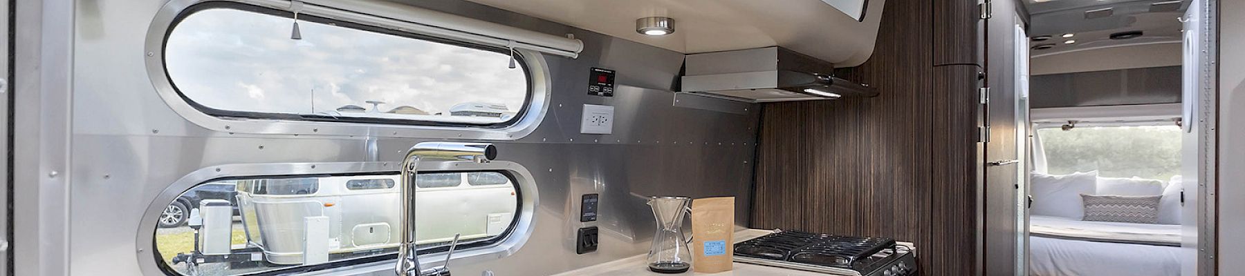 The image shows a modern RV interior with a kitchen, featuring a sink, stove, and cabinets, leading to a bed in the background.