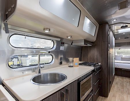 The image shows a modern RV interior with a kitchen, featuring a sink, stove, and cabinets, leading to a bed in the background.