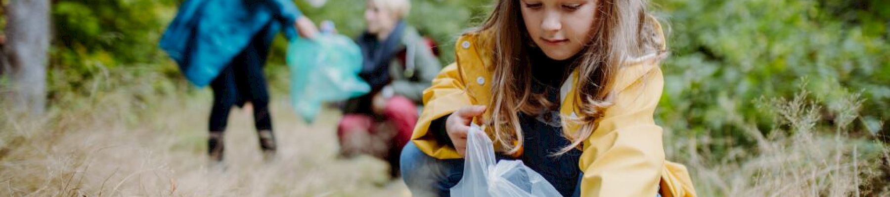 A child in a yellow raincoat picks up litter in a forest, with two adults in the background also collecting trash, surrounded by greenery.