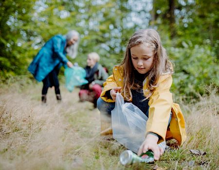 A child in a yellow raincoat picks up litter in a forest, with two adults in the background also collecting trash, surrounded by greenery.