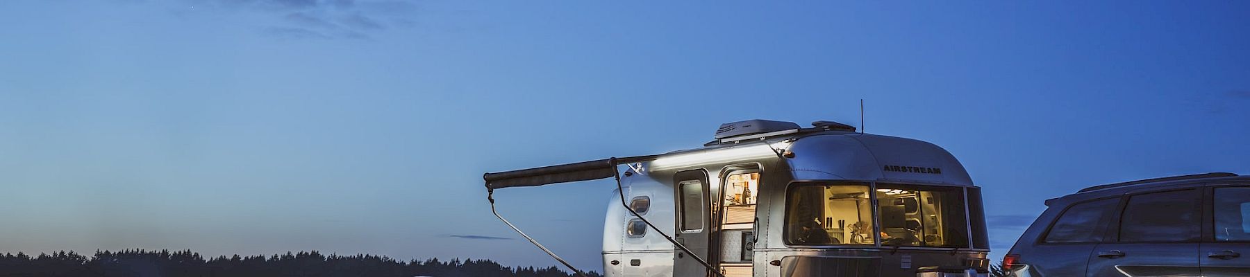 Airstream trailer and SUV parked by a lakeside at dusk, with a campfire and picnic table, under a cloudy sky.