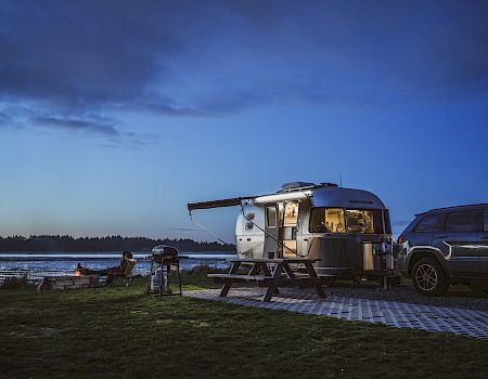 Airstream trailer and SUV parked by a lakeside at dusk, with a campfire and picnic table, under a cloudy sky.