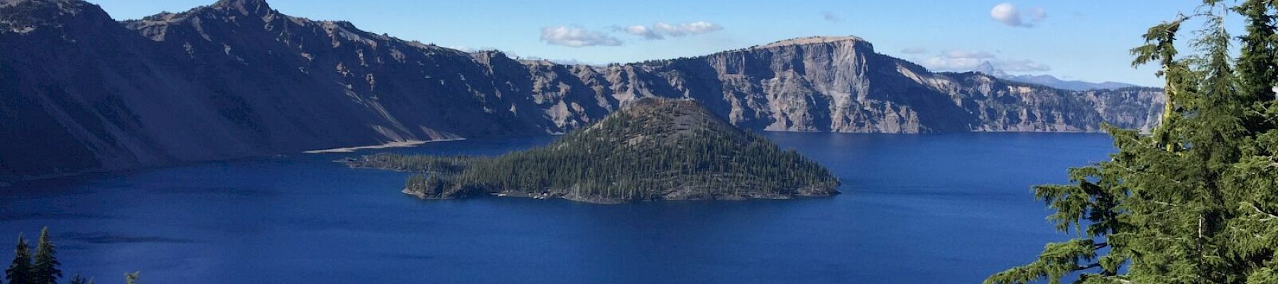 A deep blue lake surrounded by rugged mountains and dense forest, with a small island at its center under a clear blue sky.