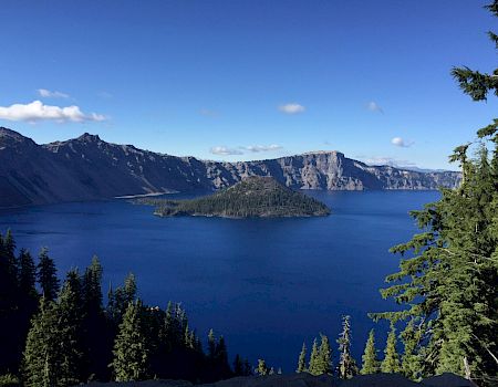 A deep blue lake surrounded by rugged mountains and dense forest, with a small island at its center under a clear blue sky.