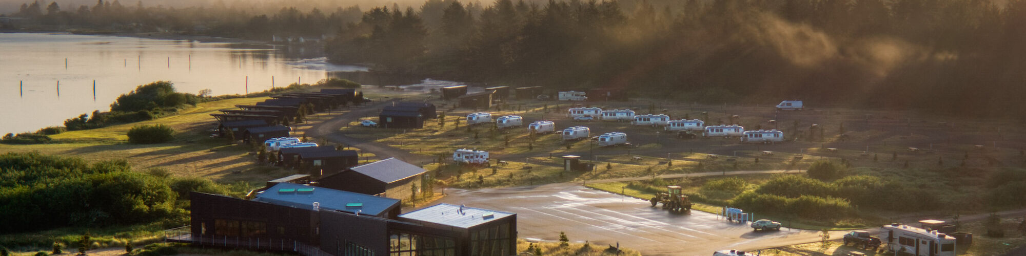 A scenic campground near a body of water at sunset, with RVs, buildings, and a forest in the background.