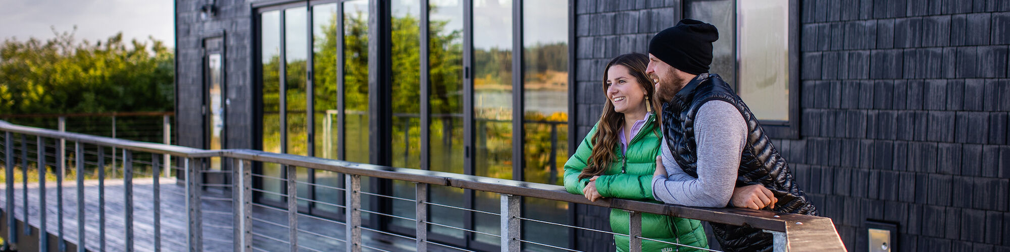 A couple stands on a deck, leaning on the railing, looking out at the view beside a modern building with large windows.