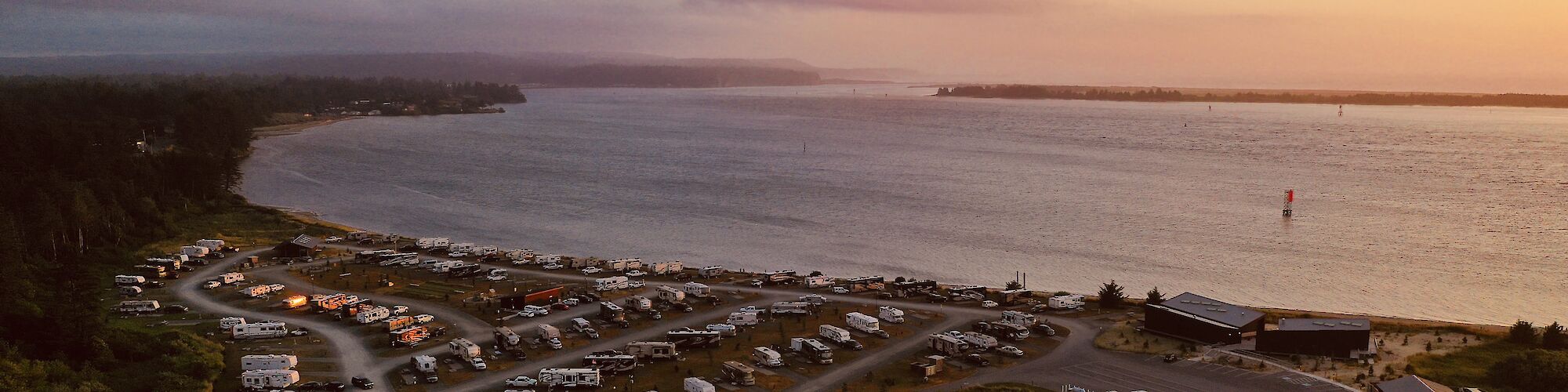 The image shows a coastal RV park with numerous campers and trailers near the shoreline during sunset, offering a peaceful and scenic view.