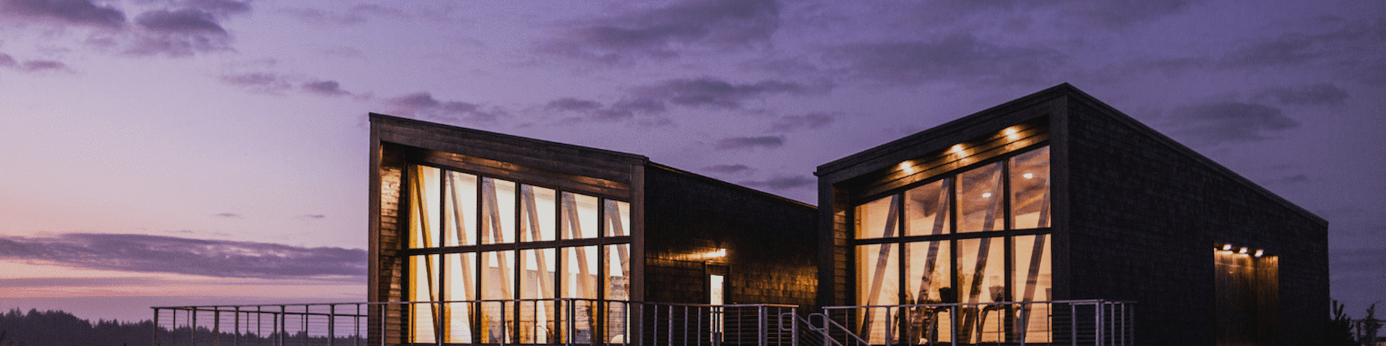 A modern building with large glass windows and wooden exterior is illuminated against a twilight sky.