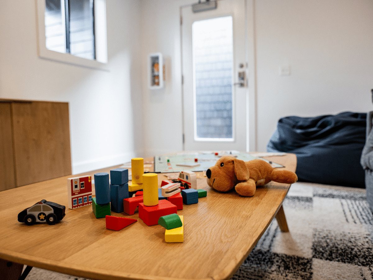 A room with a wooden table featuring toy blocks, a stuffed animal, and a toy car, with a couch and beanbag in the background.