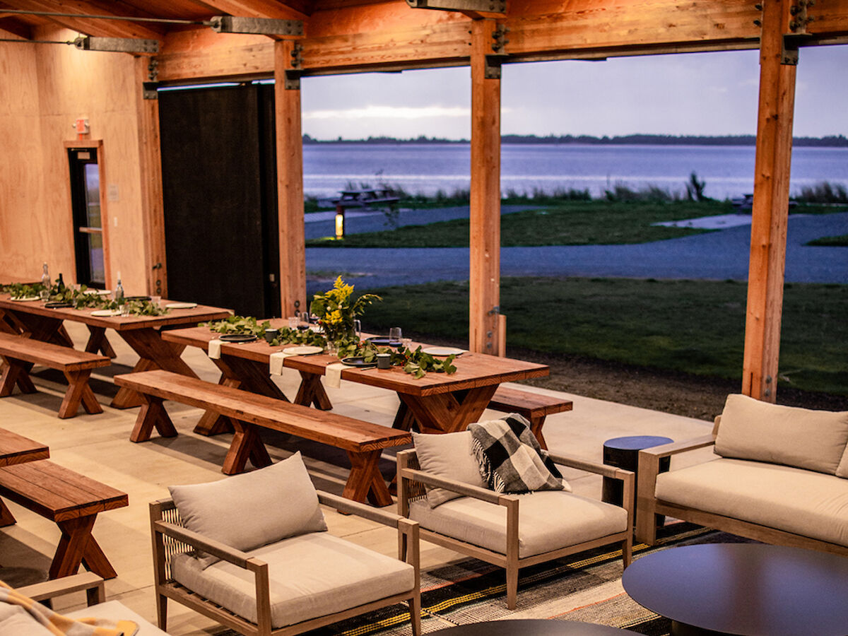 Outdoor seating area with cushioned benches and tables under a wooden pergola, overlooking a body of water at dusk.