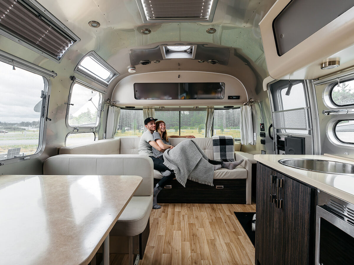 The image shows a cozy interior of an airstream suite with a couple sitting on a couch, under a blanket, surrounded by modern furnishings.