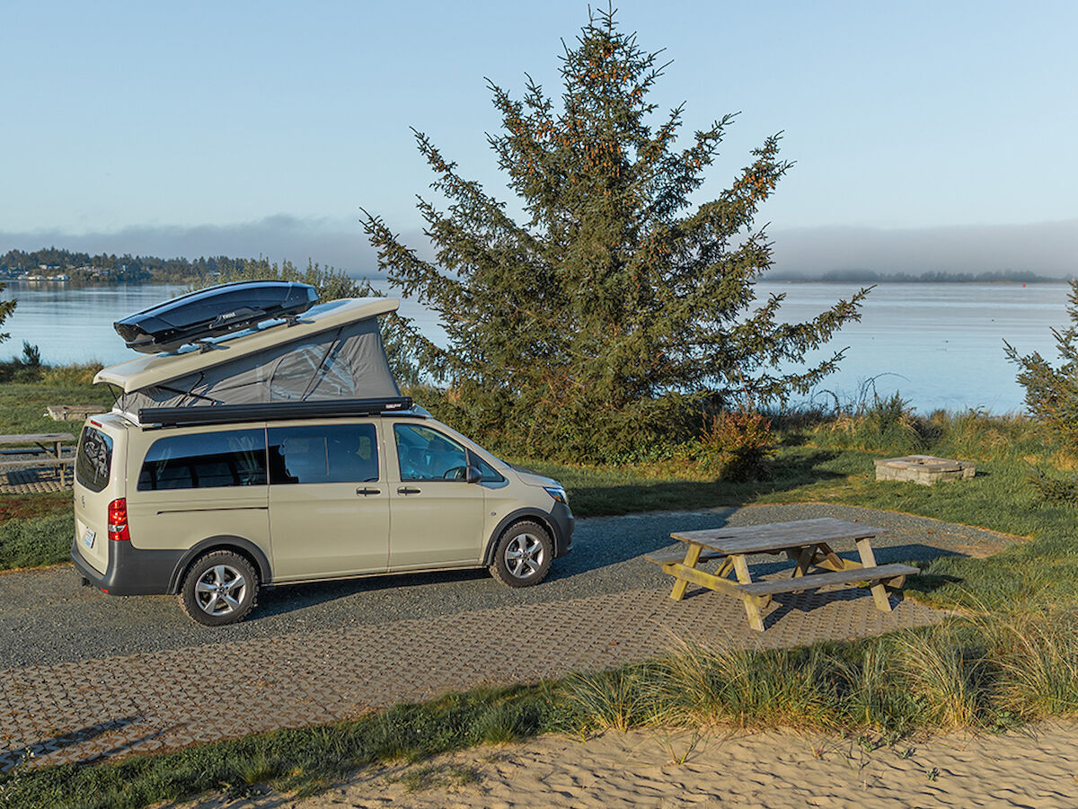 A camper van parked near a lake, with a pine tree and a picnic table in the background, surrounded by sand and grass.