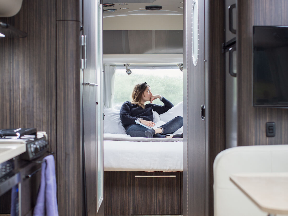 The image shows the interior of a modern camper van, featuring a kitchen, seating area, and a person relaxing on a bed in the back.