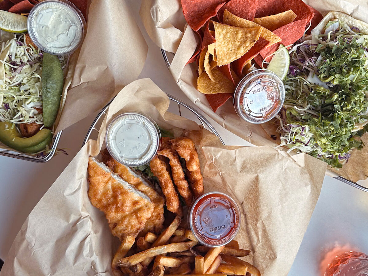 The image shows loaded nachos, fried fish with fries, sauces, and guacamole. Everything is served on paper-lined trays on a table.