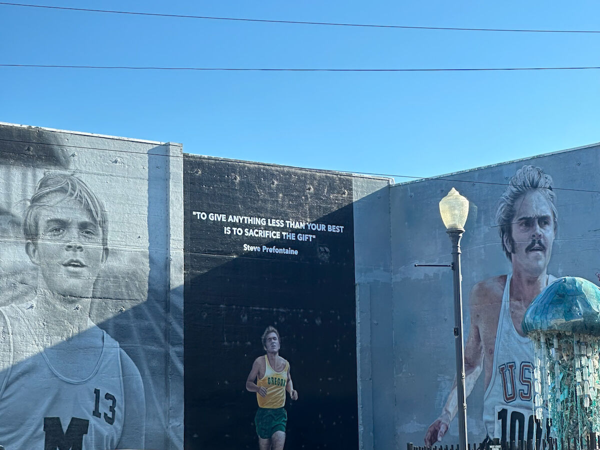 The image shows a mural with athletes and a quote. The background is a clear blue sky, and there's a streetlamp in front.